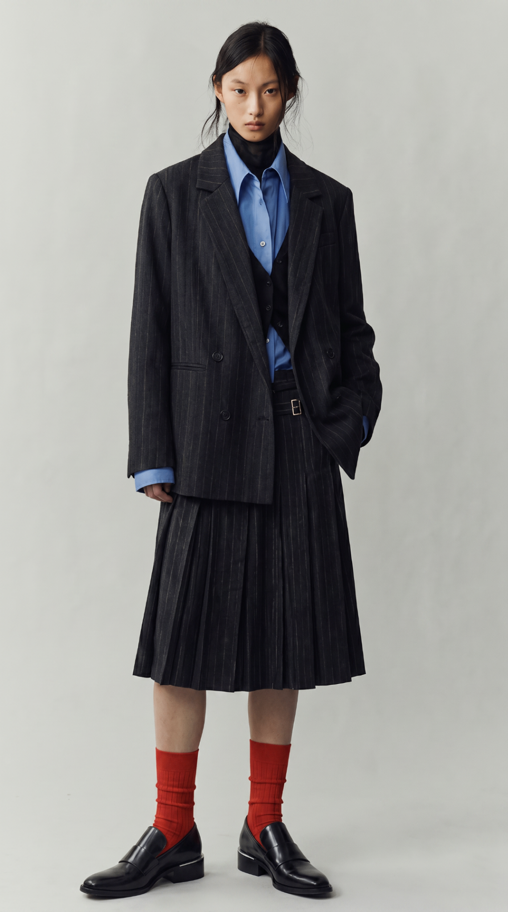 Look 01 — charcoal pinstripe blazer and pleated skirt layered over a blue shirt with bright red socks and black loafers.